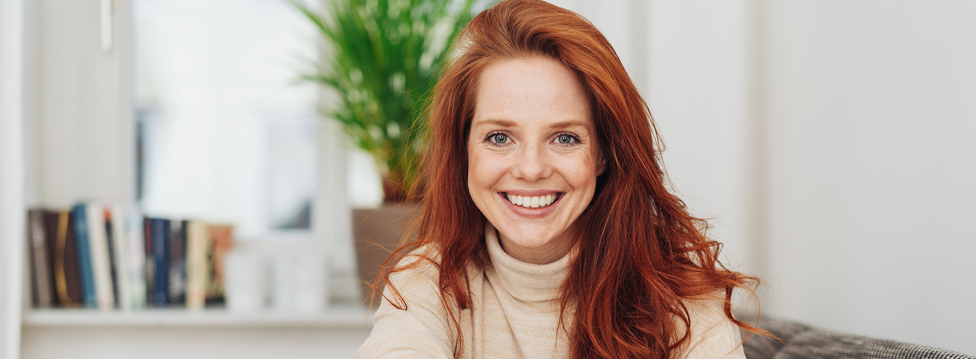 The image features a woman with red hair, wearing a brown turtleneck sweater, sitting in front of a bookshelf. She is smiling and appears to be posing for the photo.