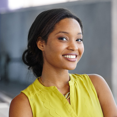 The image shows a smiling woman with dark hair, wearing a yellow top and a light-colored necklace, standing against a blurred background that appears to be an urban setting.