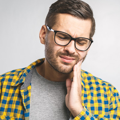 A man with a beard, wearing glasses and a yellow plaid shirt, is holding his hand to his mouth in pain or discomfort.