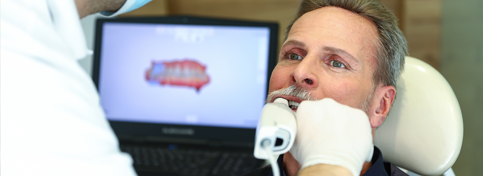 The image depicts a man receiving dental care, with a dental professional using a computer screen to guide the procedure.
