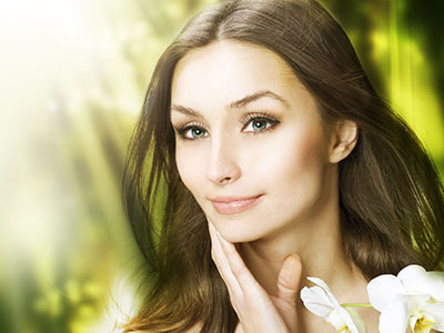 A woman with fair skin, wearing makeup and a light-colored top, is the central figure in this image. She has long hair and is gazing off to her left. The background features blurred greenery and a soft, natural lighting effect.