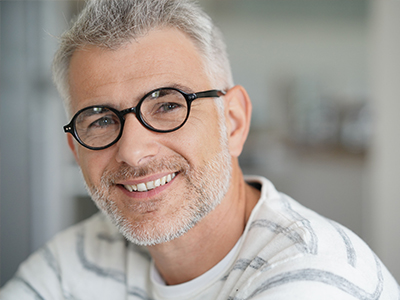 The image features a man with glasses, smiling and looking directly at the camera. He has short hair, a beard, and is wearing a patterned shirt.