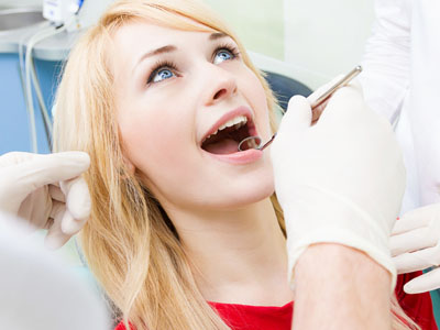 A woman is seated in a dental chair, receiving dental care with her mouth open and a dental professional working on her.