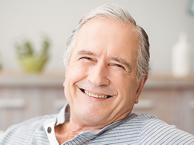 The image shows an older man with gray hair, smiling and looking directly at the camera. He is seated comfortably in a chair, wearing a blue shirt and appears to be indoors, possibly in a living room or similar space.